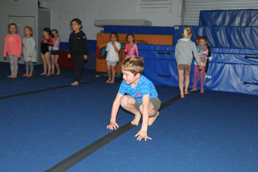 Children in a gymnasium, some standing and others moving around. One child in the foreground is crouching on the blue padded floor. Gym mats and equipment are visible in the background.