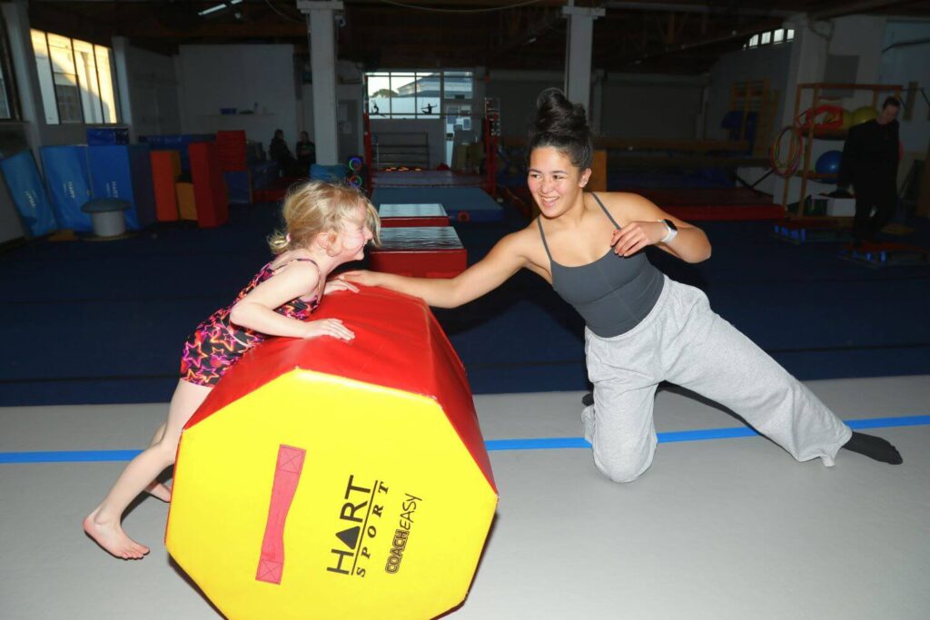 A young girl in a gymnastics outfit plays with a large, padded shape while a smiling female coach assists her on a gym floor.