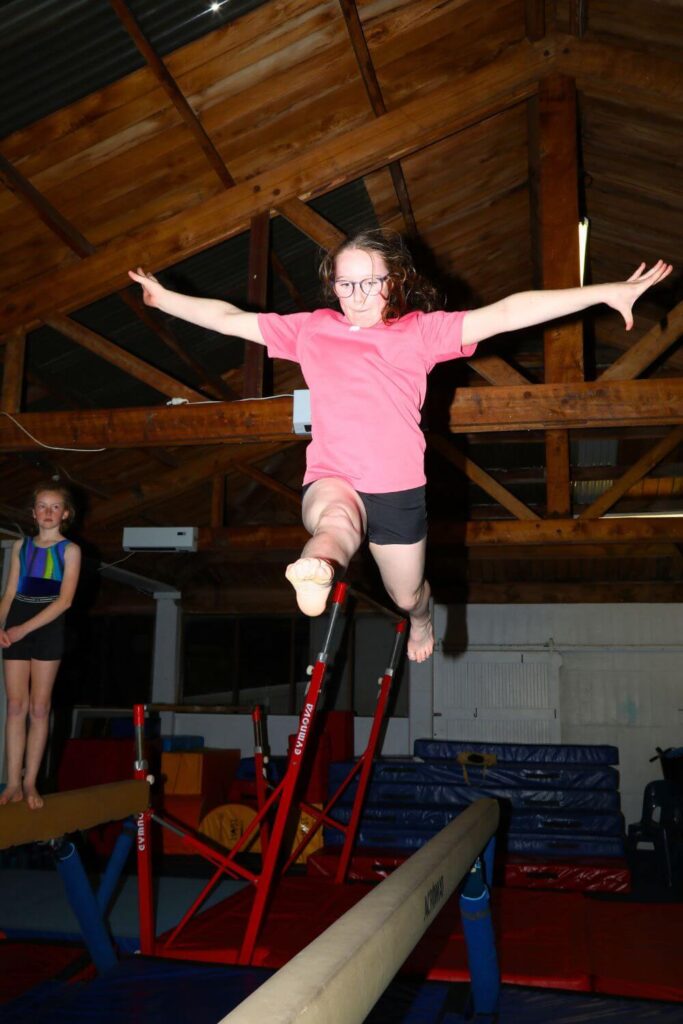 A girl in a pink shirt and black shorts practices a leap on a balance beam in a gymnasium, with another girl standing on a balance beam in the background.