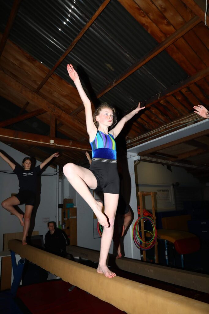 A young gymnast in a blue leotard balances on one leg with arms raised on a balance beam in a gym, while others practice in the background.