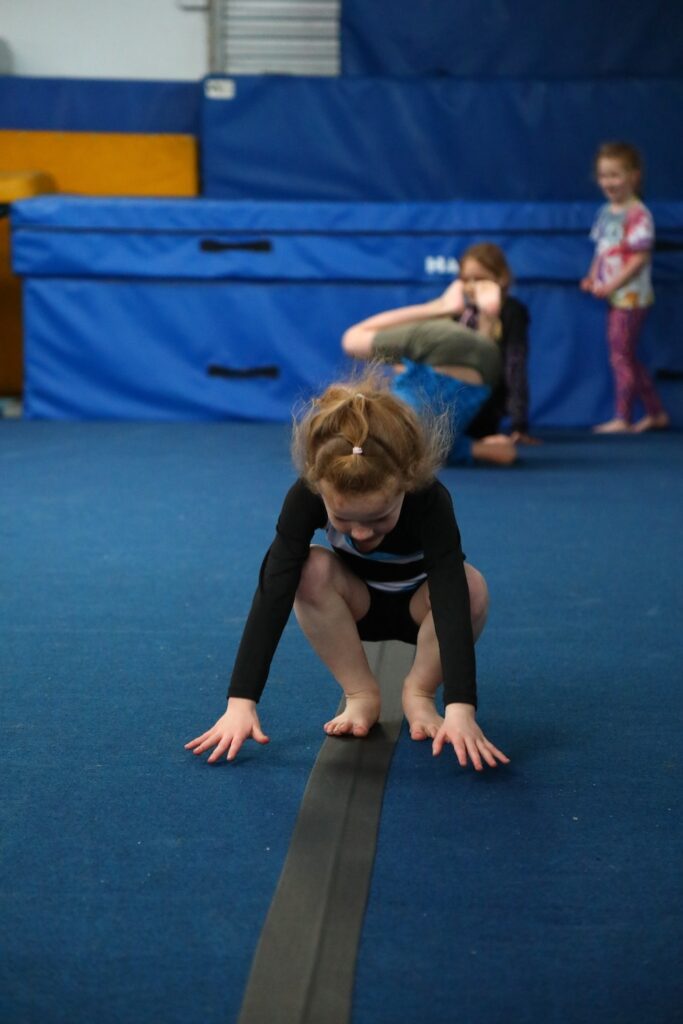 A child in a gymnastics outfit balances on a floor beam, while two other children play in the background on blue mats.