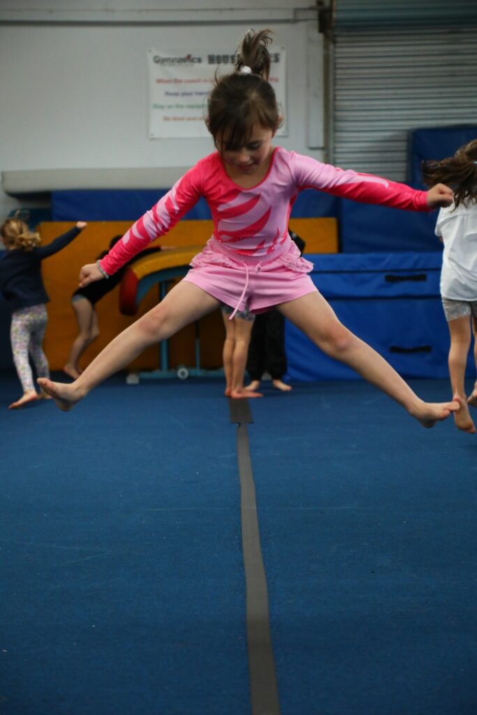 A young girl in a pink outfit performs a split jump on a balance beam in an indoor gym, with other children visible in the background.