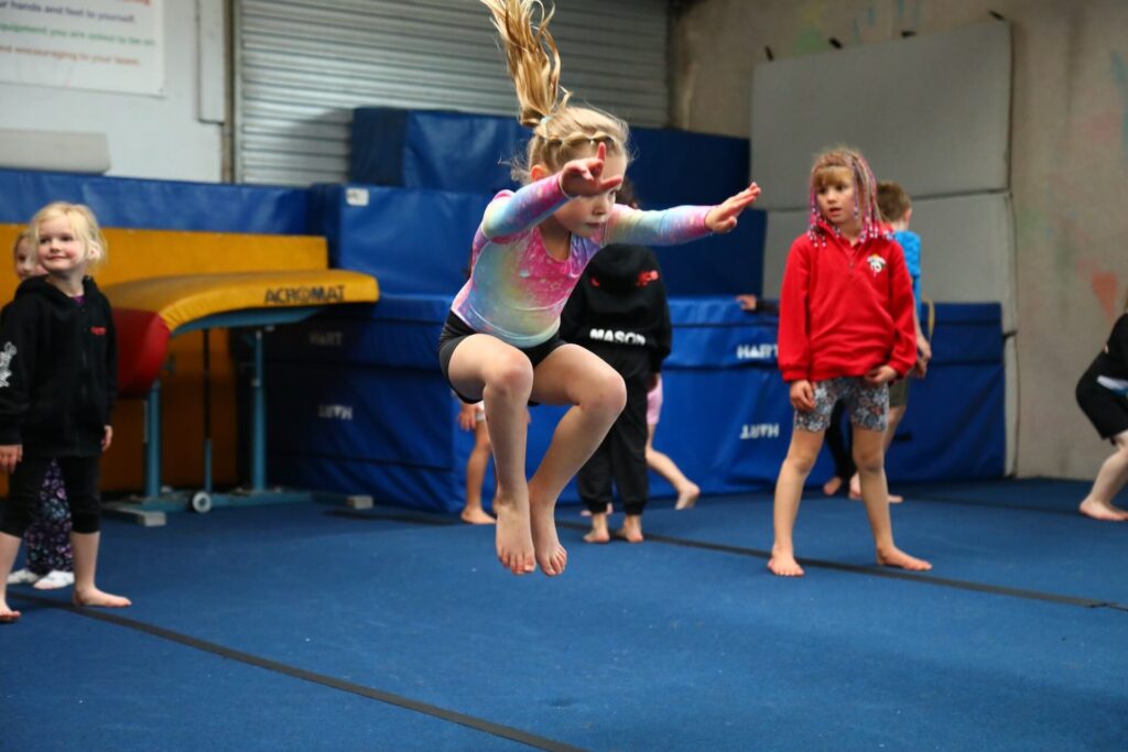 A young girl wearing a colorful leotard jumps into the air in a gymnasium, with other children watching.