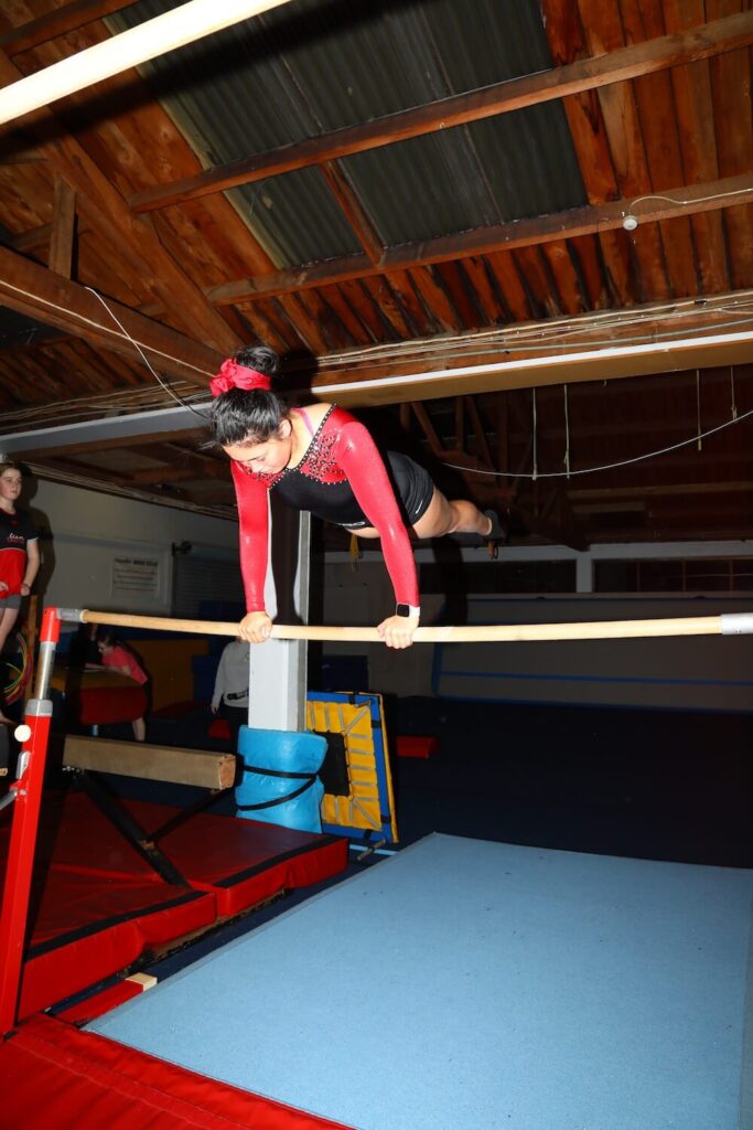 A gymnast in a red and black leotard performs a horizontal bar routine in an indoor gym.