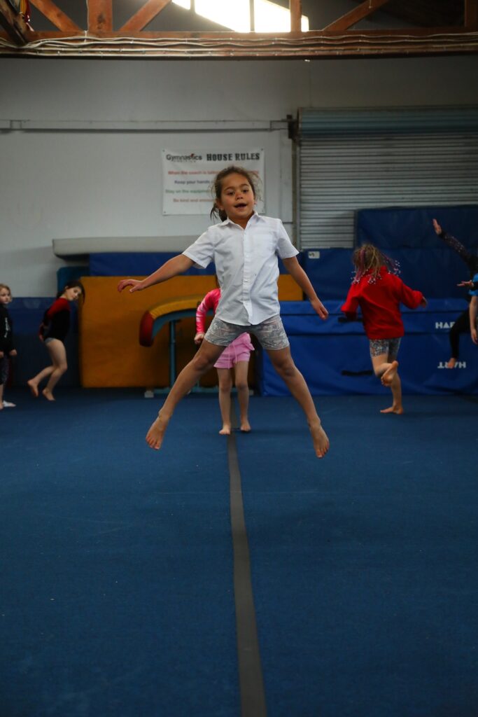 A child is mid-air while jumping in a gymnasium with other children and gym equipment in the background.