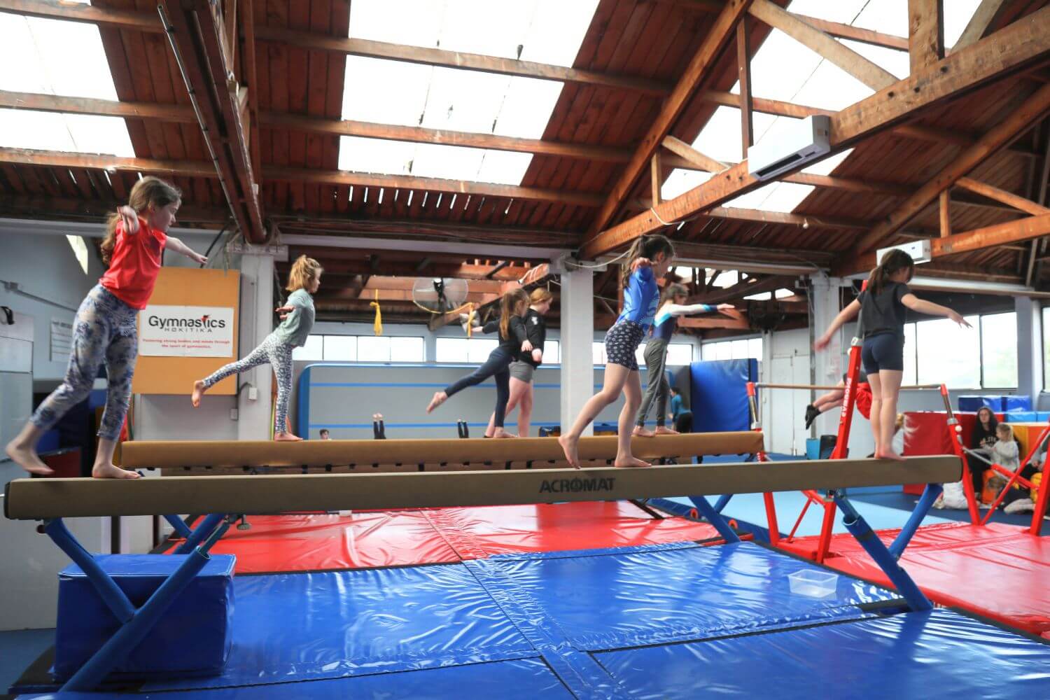 Young gymnasts practice balancing on beams in a spacious, well-lit gymnasium with high wooden ceilings.