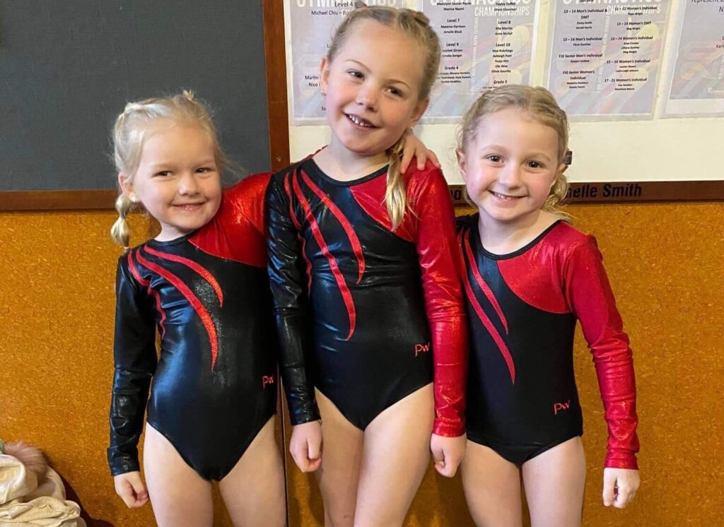 Three young girls in matching black and red gymnastics leotards stand together, smiling at the camera. They have blonde hair styled in pigtails and are standing in front of a bulletin board.