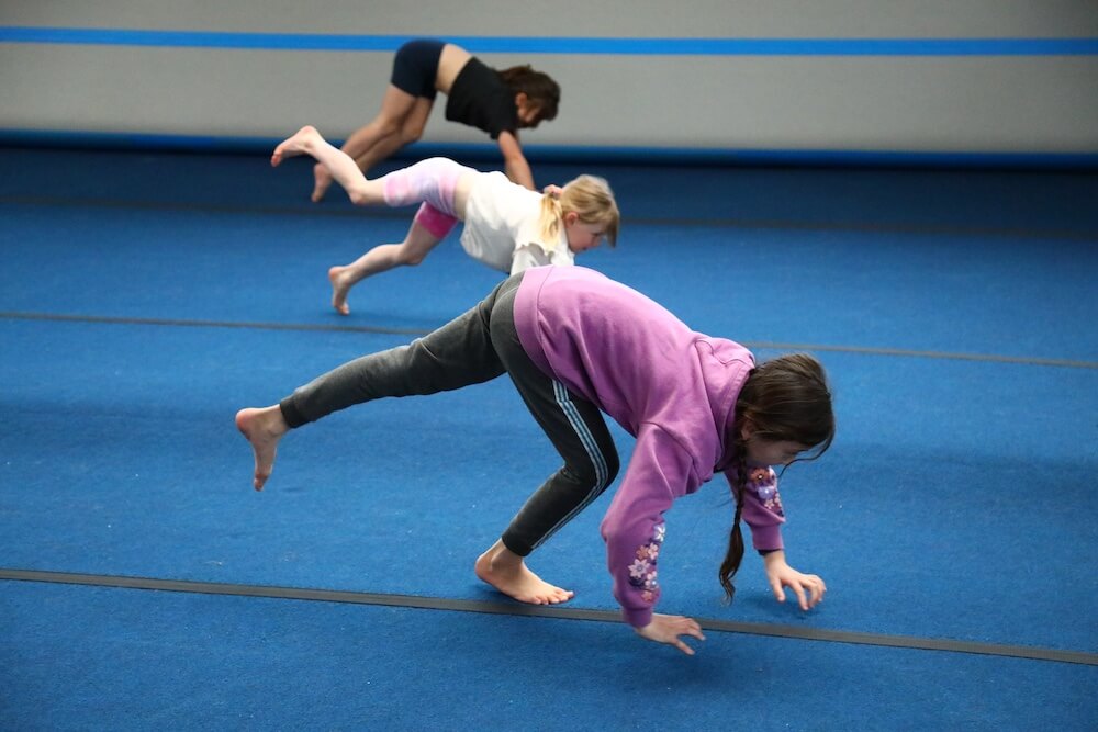 Three children are practicing bear walk exercises on a blue gym mat, with one leading and the others closely following.