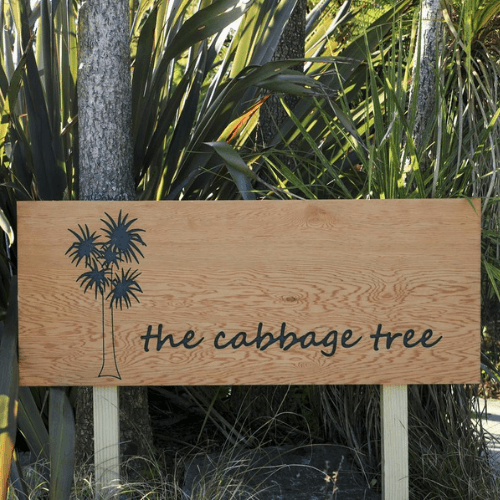 Wooden sign with the text "the cabbage tree" and a silhouette of a tree, surrounded by foliage.