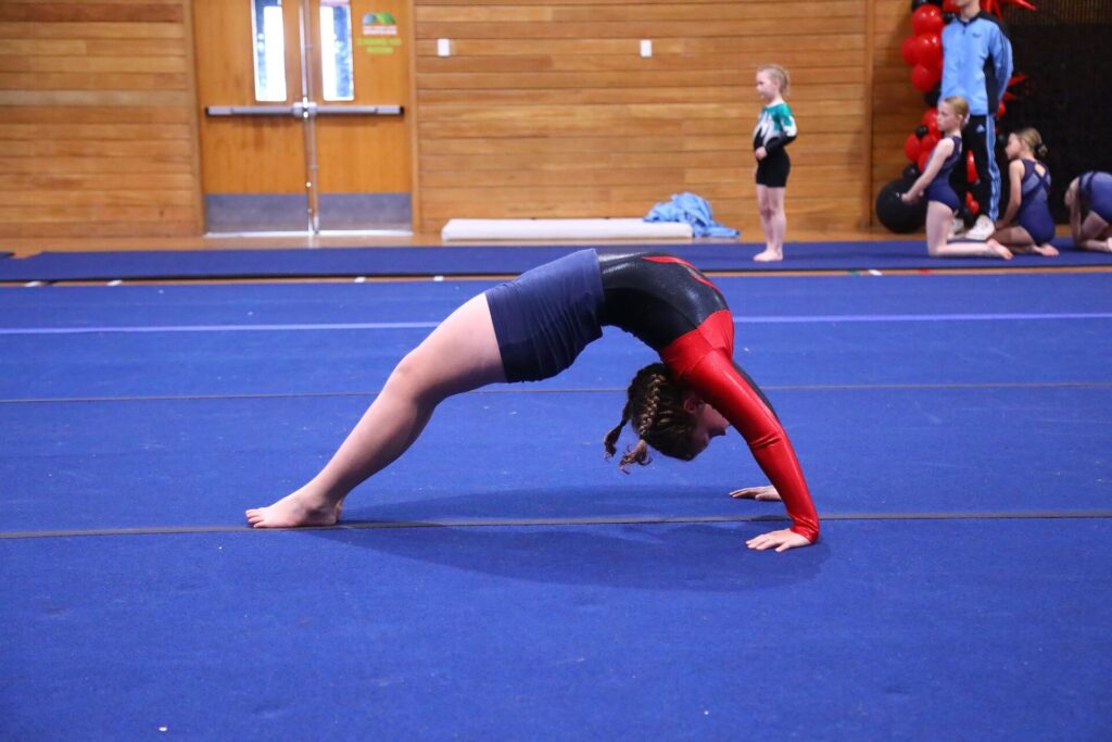 A girl in a gymnastics uniform performs a backbend on a blue mat inside a gymnasium. Other gymnasts and balloons are visible in the background.