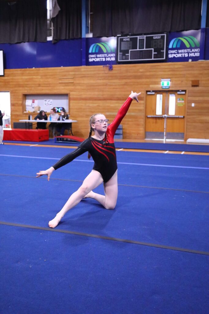 A gymnast in a red and black leotard performs a floor routine on a blue mat in a sports hall.