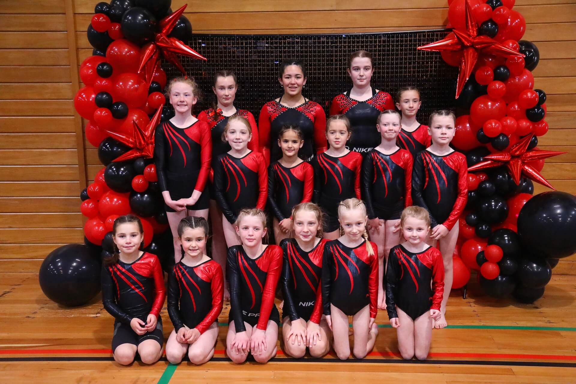 A group of gymnasts in matching red and black uniforms poses in front of a balloon arch.