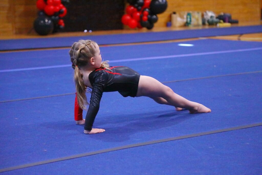 A young gymnast performs a bridge position on a blue mat, wearing a black and red leotard. Red and black balloons are in the background.