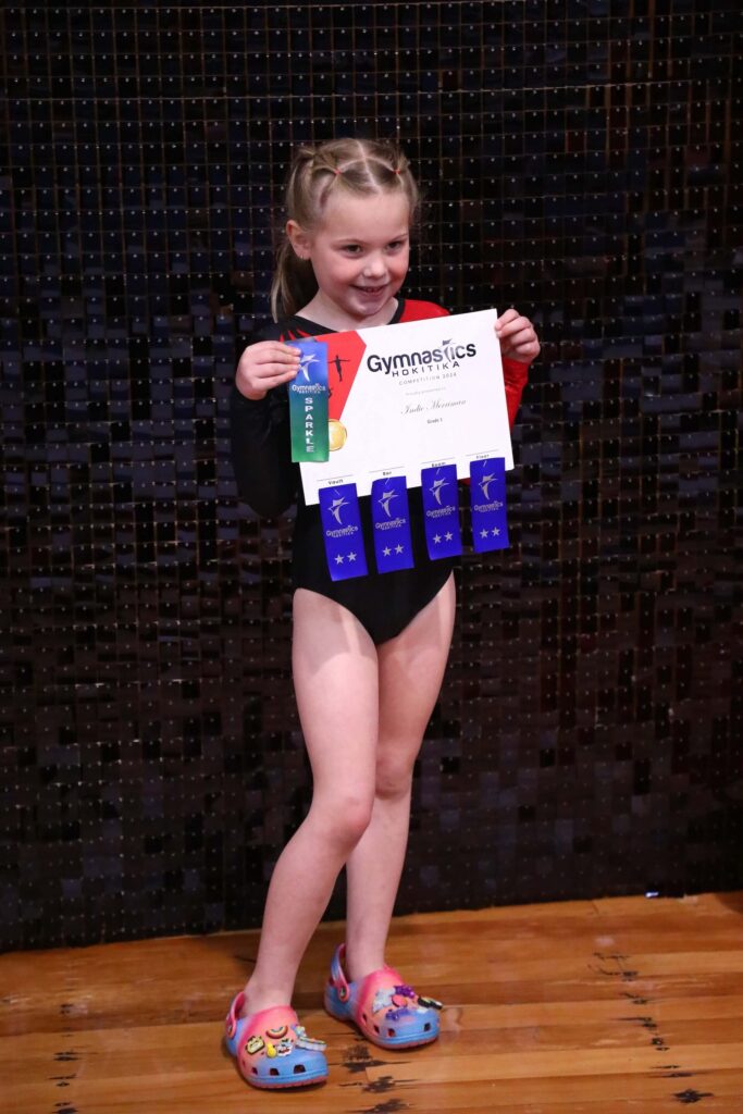 A young girl in a leotard holds a gymnastics certificate and multiple award ribbons, standing on a wooden floor.