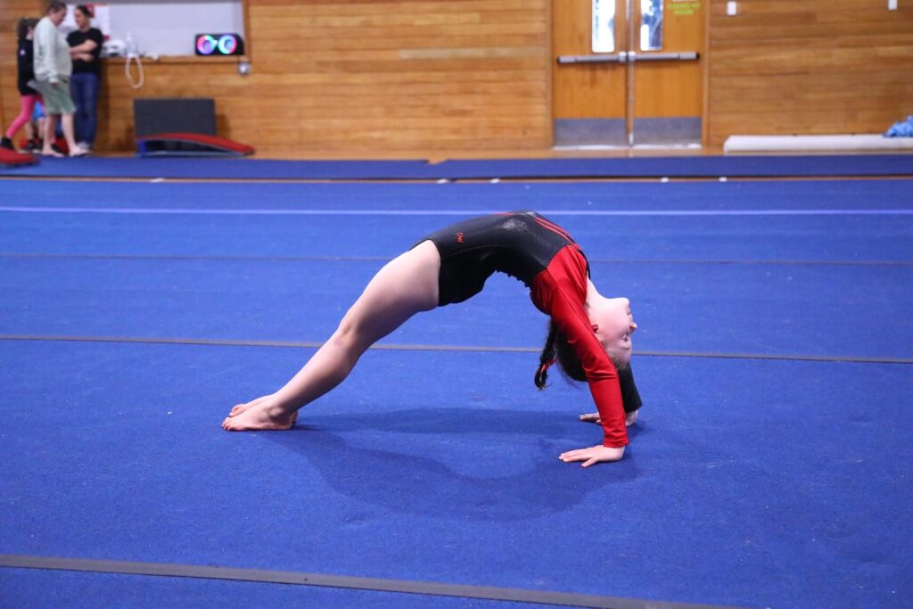 A gymnast in a red and black leotard performs a backbend on a blue mat in a gymnasium.