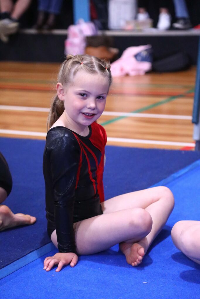 A young girl in a gymnastics outfit sits on a blue mat, smiling at the camera.