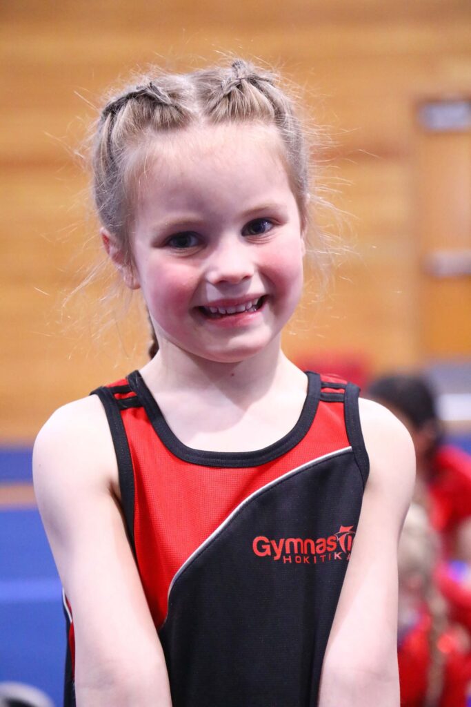 Young girl with braided hair wearing a black and red gymnast outfit smiles at the camera indoors.