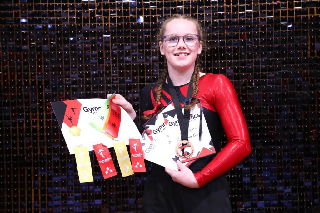 Young gymnast in a red and black outfit holds certificates, ribbons, and a medal, smiling in front of a sparkly backdrop.