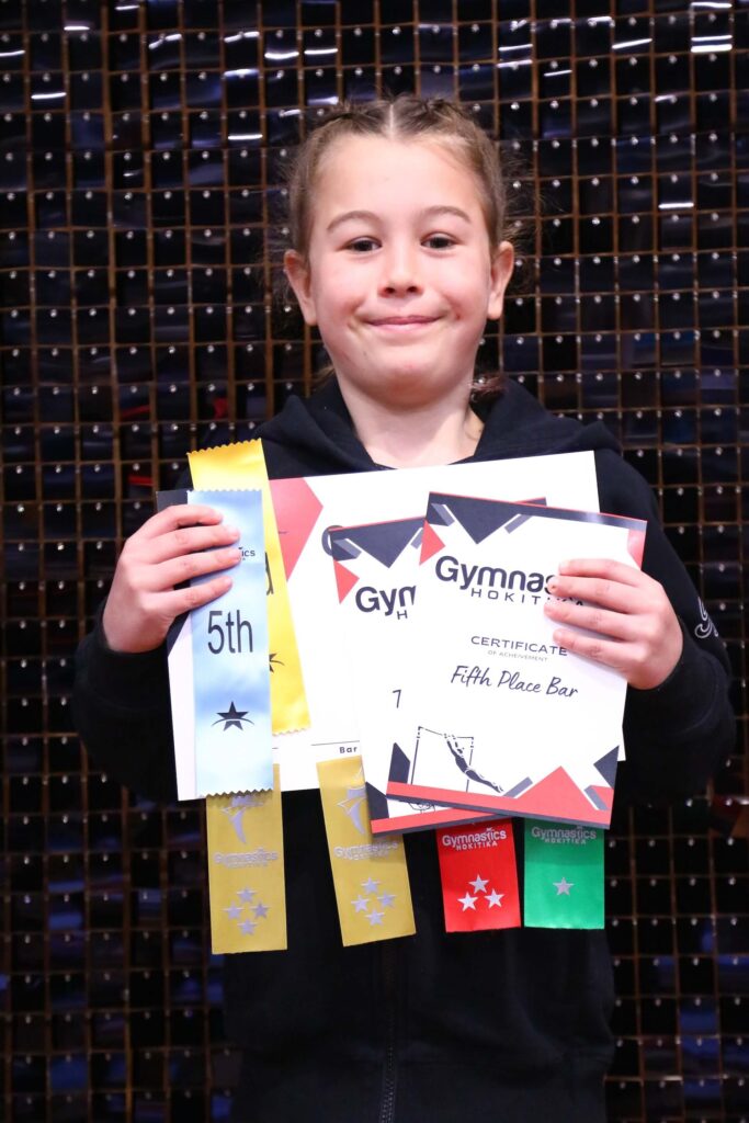 A young girl smiling, holding five gymnastics certificates and ribbons, standing against a tiled wall.