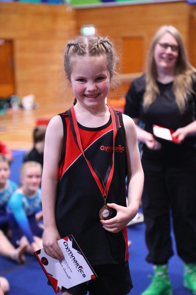 A young girl with braids smiles while holding a gymnastics certificate and wearing a medal. She stands in a gymnasium with other people in the background.