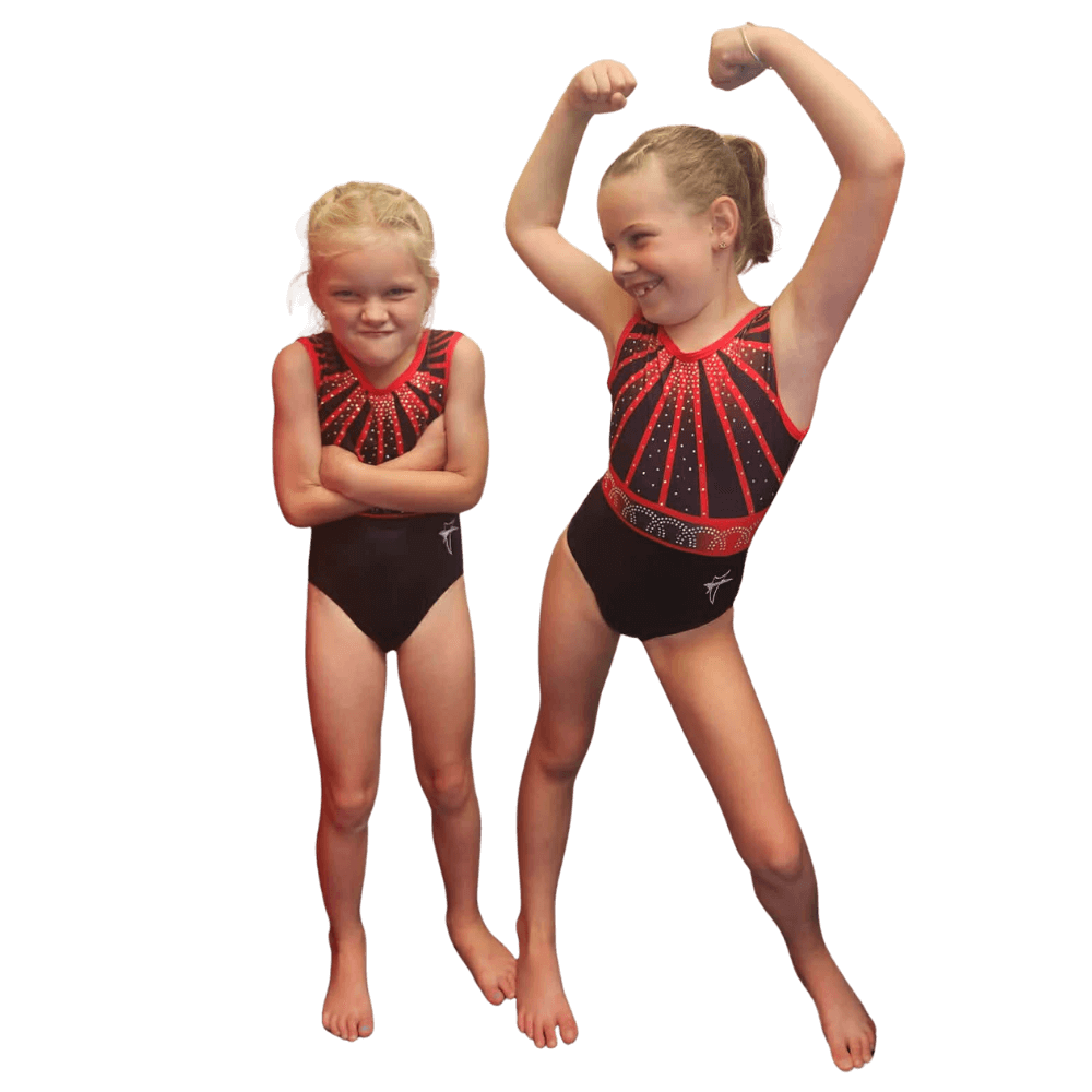 Two young girls wearing matching black and red gymnastics leotards stand barefoot, one with arms crossed and the other flexing her muscles.