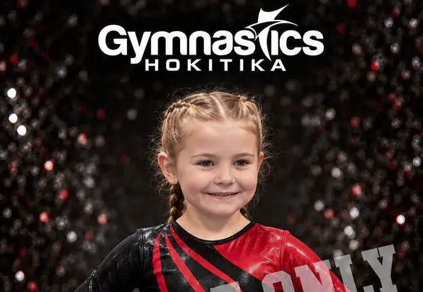 A young gymnast in a red and black leotard smiling in front of a Gymnastics Hokitika logo.